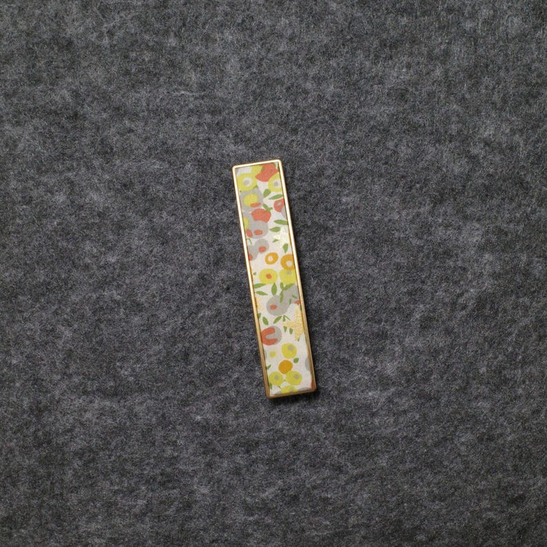 May include: A rectangular tie bar featuring a gold-toned metal frame and a decorative inlay. The inlay displays a repeating pattern of stylized flowers and leaves in shades of orange, yellow, light green, and grey against a white background.