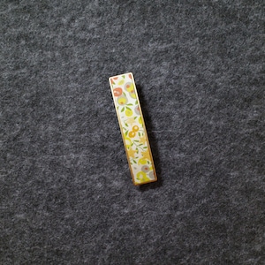 May include: A rectangular tie bar featuring a gold-toned metal frame and a decorative inlay. The inlay displays a repeating pattern of stylized flowers and leaves in shades of orange, yellow, light green, and grey against a white background.
