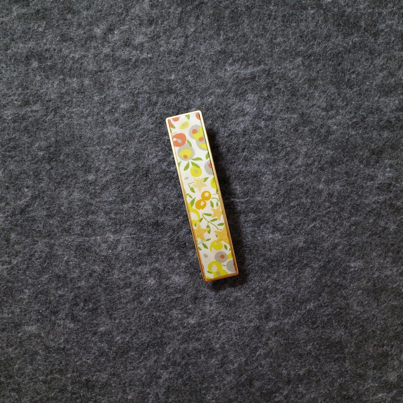 May include: A rectangular tie bar featuring a gold-toned metal frame and a decorative inlay. The inlay displays a repeating pattern of stylized flowers and leaves in shades of orange, yellow, light green, and grey against a white background.