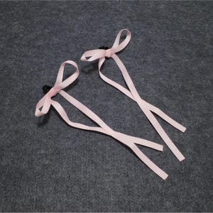 May include: Two pink ribbon hair clips with black clasps. The ribbons are tied in bows.