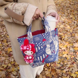 Peut inclure: Un sac fourre-tout bleu et blanc avec une doublure rouge et un motif floral. Un jouet en peluche rouge avec un nœud rose est attaché au sac. Un gobelet à emporter blanc est dans le sac.