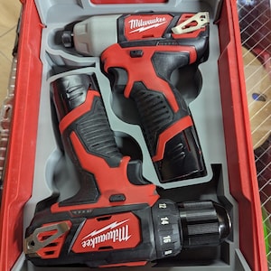 May include: A red and black Milwaukee power tool set in a red plastic case. The set includes a drill and an impact driver. The case has a gray interior with molded compartments to hold the tools securely. The Milwaukee logo is visible on each tool.