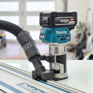 Könnte beinhalten: Ein kabelloser Makita-Fräser mit einem schwarzen und türkisfarbenen Gehäuse, der an einer Schiene befestigt ist. Der Fräser hat einen schwarzen Akku. Die Schiene ist silberfarben mit dem Makita-Logo.