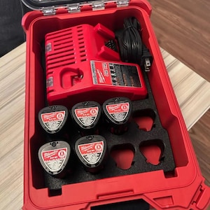 May include: A red Milwaukee power tool kit with a charger and multiple batteries. The kit is open, revealing the charger, batteries, and a black foam insert. The batteries are black with red and white labels.