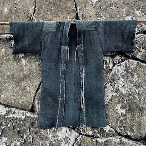 Puede incluir: Una túnica japonesa "Jinbei" de color índigo oscuro, una prenda tradicional, colgada de una barra de madera sobre un fondo de pared de piedra.