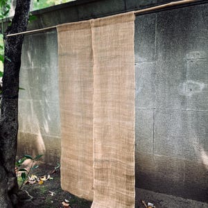 Op de afbeelding: Een paar rechthoekige, lichtbruine, geweven stoffen panelen die verticaal aan een dunne, horizontale houten roede hangen. De stof heeft een zichtbaar, grof, getextureerd weefsel en een natuurlijke, aardse kleur, gelijkmatig opgehangen in twee delen.