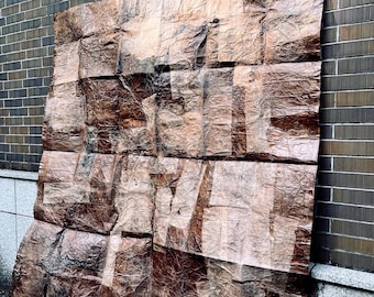 May include: A large, rectangular artwork with a crumpled, textured surface. The piece is predominantly brown with lighter patches, possibly displaying text. It's leaning against a brick wall, suggesting an outdoor installation or display.