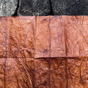 May include: A rectangular piece of brown, textured paper, possibly handmade, is suspended horizontally. The paper has a wood-grain appearance with visible creases and folds. It is held by a thin, light-colored wooden dowel against a dark, textured stone background.
