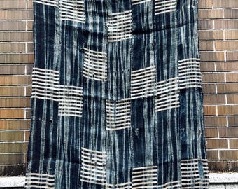 May include: A dark blue and white patterned textile hanging against a brick wall. The textile features a grid-like design with vertical stripes and horizontal blocks of white lines, creating a textured appearance. The fabric appears to be handmade.