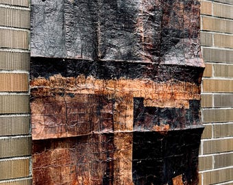 May include: A square wall art piece featuring a heavily textured, crinkled surface with a dark color palette of deep black and burnt orange. The material appears layered and weathered, displaying a grid-like pattern of creases and folds.