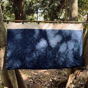 Op de afbeelding: Een blauwe en witte stof met een patroon hangt aan een houten stok tussen twee bomen. De stof heeft een geometrisch ontwerp met een witte rand.