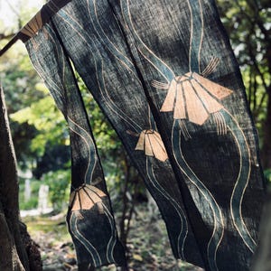 以下が含まれることがあります： 紺色の生地に、様式化されたベージュの花の形と流れるようなラインが繰り返されるパターン。 生地は屋外に吊るされており、太陽の光が素材を透過し、柔らかく自然な美しさを作り出しています。