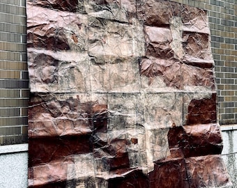 May include: A large, rectangular artwork with a textured, crumpled appearance. The piece is primarily brown with lighter beige and darker reddish-brown sections, creating a layered effect. It is mounted on a brick wall.