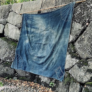 May include: A large, blue denim fabric panel hangs against a stone wall. The fabric has a faded, worn look and is decorated with white stitching in a geometric pattern.