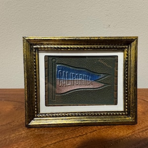 May include: A framed vintage nautical flag with the word "CALIFORNIA" in blue and red. The flag is set against a textured green background, framed in a gold-colored, ornate frame. The frame is rectangular and sits on a wooden surface.
