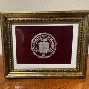 May include: A rectangular gold-toned frame with a beaded inner border containing a maroon velvet background. Centered on the velvet is a circular silver-coloured University of Minnesota emblem featuring the university seal and text in relief.