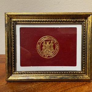 May include: A framed emblem featuring the Massachusetts Institute of Technology seal. The seal is gold, set against a burgundy background, and framed in a gold-coloured, ornate frame. The frame is rectangular and sits on a wooden surface.