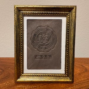 Peut inclure: Un cadre rectangulaire doré avec une bordure intérieure perlée contenant un insert en cuir gris foncé. L'insert présente un sceau circulaire en relief du Massachusetts Institute of Technology et les lettres M.I.T. imprimées sous le sceau.