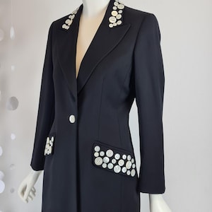 Pode incluir: Um blazer preto com botões brancos decorativos na gola, bolsos e frente. Os botões redondos têm um brilho perolado. O blazer é exibido em um manequim. O design é clássico e sob medida.