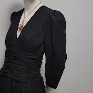 以下が含まれることがあります： Vネックと長袖の黒いドレスがマネキンに展示されています。ドレスはシャーリングウエストと花柄が特徴です。ピンクのペンダントが付いたシルバーのネックレスがネックラインを飾っています。マネキンは白い背景にあります。