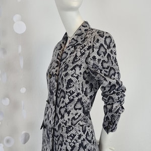 Peut inclure: Un blazer gris et noir à motif peau de serpent présenté sur un mannequin. Le blazer a un col cranté, des boutons et des manches longues. Le tissu a un aspect texturé et le style est ajusté.