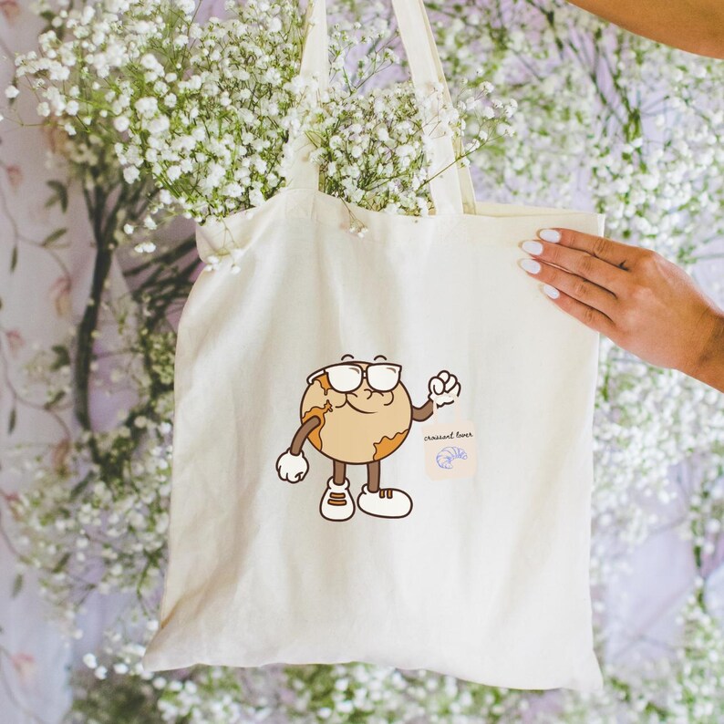 May include: Cream-colored tote bag featuring a cartoon earth character wearing glasses and sneakers. The earth character is brown and tan with white gloves and shoes. The bag has a small blue graphic and the words "crescent lover".