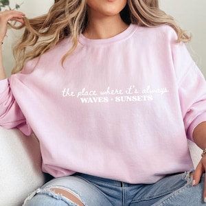 May include: A light pink sweatshirt with the text "the place where it's always WAVES + SUNSETS" in white script. The sweatshirt is a solid color and has a crew neck.