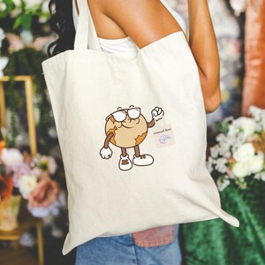 May include: Cream-colored tote bag featuring a cartoon earth character wearing sunglasses and sneakers. The earth character is holding up a fist. A small square graphic with the words "croissant tour" is on the bag.