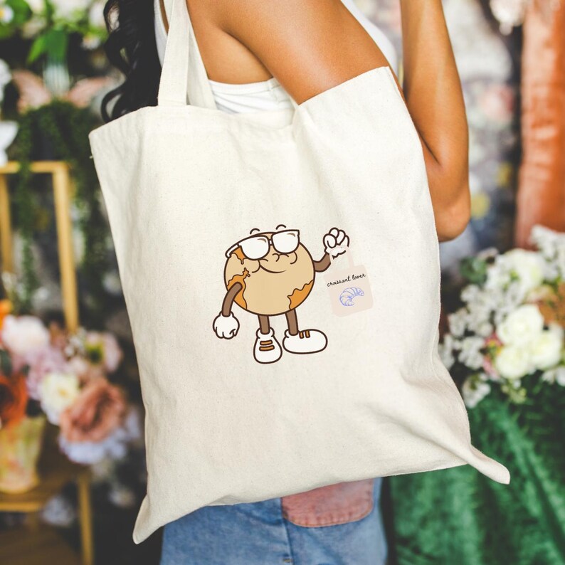 May include: Cream-colored tote bag featuring a cartoon earth character wearing sunglasses and sneakers. The earth character is holding up a fist. A small square graphic with the words "croissant tour" is on the bag.