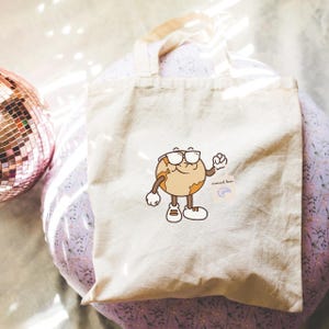 May include: A beige canvas tote bag featuring a cartoon character resembling a planet wearing glasses and sneakers. The character is brown and tan with a smiling face. The bag has a small tag with the text "crescent lover".