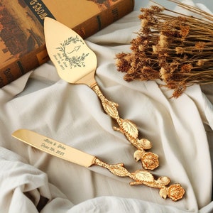 May include: Gold wedding cake knife and server set with floral designs and personalised engraving. The knife has the text "Alicia & John June 7th, 2023" and the server has the text "A & J June 7th, 2023".