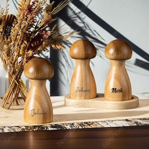 May include: Three wooden mushroom-shaped salt and pepper grinders. The grinders are light brown with a darker brown cap. The names "Johan" and "Mark" are printed on two of the grinders. The grinders sit on a wooden tray.