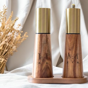 May include: A set of two wooden salt and pepper shakers with gold metal tops. The shakers are engraved with the initials "H" and "M" with floral designs and the names "Herbert" and "Mia".