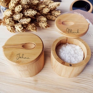 Peut inclure: Deux salières en bambou personnalisées avec des cuillères en bois. Une salière est ouverte et remplie de sel blanc. Les salières sont étiquetées "Johan" et "Mark".