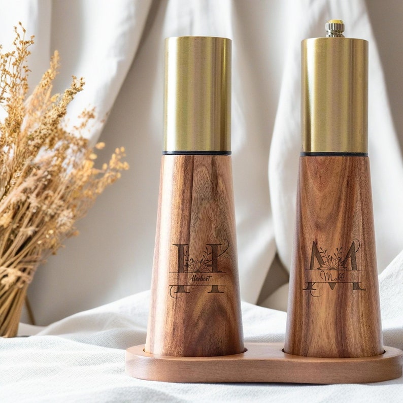 May include: A set of two wooden salt and pepper shakers with gold tops. The shakers are engraved with the letters "H" and "M" with floral designs and the names "Herbert" and "Mary" underneath. The shakers are on a wooden base.