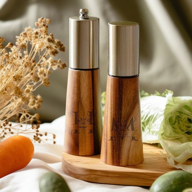 May include: A set of two wooden salt and pepper shakers with engraved initials and floral designs. The shakers are on a wooden base and have stainless steel tops.