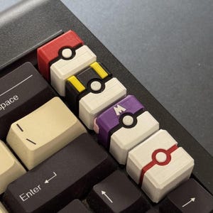 May include: Close-up of a computer keyboard with custom keycaps. The keycaps are designed to resemble various Pokeballs, featuring red, white, black, yellow, and purple colors. The keyboard keys are black and beige.