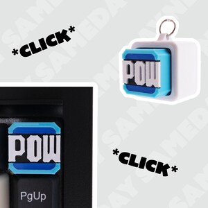 Peut inclure: Un capuchon de touche blanc et bleu avec le mot "POW" imprimé dessus. Le capuchon de touche est sur un clavier noir avec une touche blanche étiquetée "PgUp".