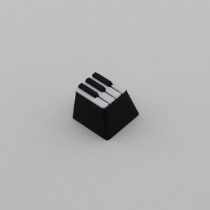 May include: A black and white keyboard keycap with a piano key design. The keycap is square shaped and has a black base with four white keys on top.