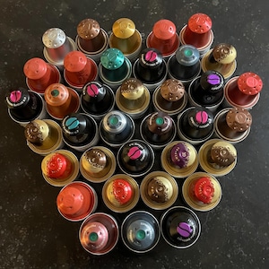 May include: A selection of espresso coffee capsules, each with a different coloured top and label. The capsules are arranged in a heart-like shape. The tops are various colours, including silver, gold, red, and brown.