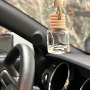 May include: A clear glass car air freshener with a wooden top and a beige string hangs in front of a car dashboard. The car interior is black, and the background is blurred.