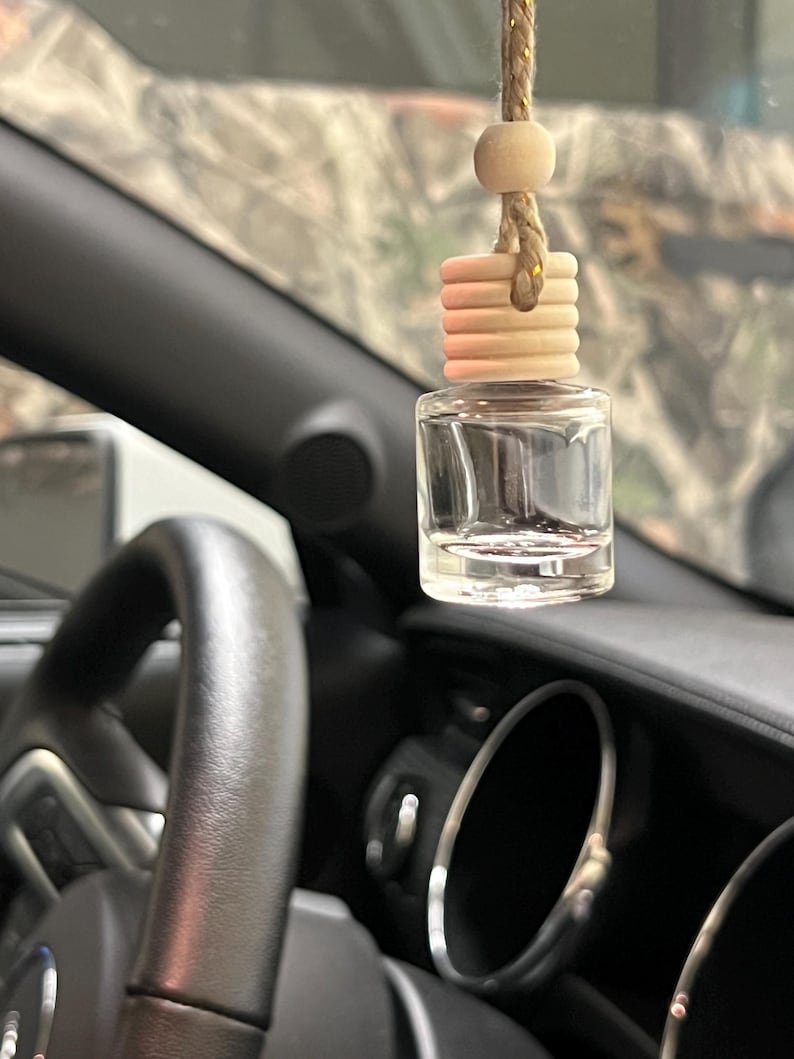 May include: A clear glass car air freshener with a wooden top and a beige string hangs in front of a car dashboard. The car interior is black, and the background is blurred.