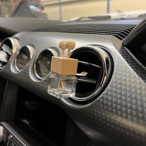 May include: A clear glass bottle with a wooden lid and two wooden sticks. The bottle is hanging from a car's air vent.