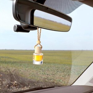 May include: A car air freshener with a small, clear glass bottle filled with yellow liquid. The bottle has a wooden cap and is suspended from the rearview mirror by a woven cord. The background shows a field and sky.