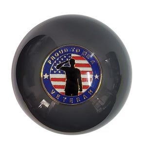 May include: A circular black object featuring a central emblem with a gold border. The emblem displays a silhouette of a person saluting against a red, white, and blue American flag background. The blue border reads PROUD TO BE A VETERAN.