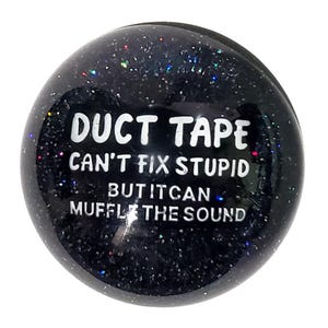 May include: A black, glitter-covered ball with white text that reads "DUCT TAPE CAN'T FIX STUPID BUT IT CAN MUFFLE THE SOUND". The ball is round and has a shiny, reflective surface.