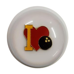 May include: A round white button featuring a central graphic design. The design includes a yellow letter I, a red heart shape, and a black bowling ball with three gold finger holes, creating a symbolic representation of a love for bowling.