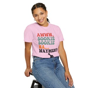 May include: A light pink t-shirt with the text "AWWW, SOOKIE SOOKIE NA... WAYMENT!" in red, green, orange, and black. The shirt is paired with blue jeans.