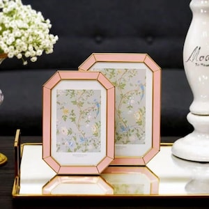 May include: Two pink picture frames with a gold trim and a floral print background. The frames are displayed on a mirrored tray with a gold edge. The frames have an octagonal shape.