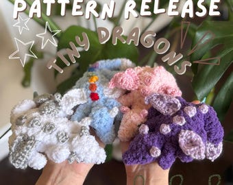 May include: Four crocheted tiny dragons in different colors: white, light blue, pink, and purple. The dragons are held in a person's hands. The text "PATTERN RELEASE TINY DRAGONS" is written in white on a black background.